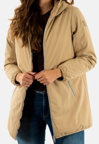 Chaqueta ligera de color beige claro con capucha, cremallera frontal y puños elásticos. Cuenta con bolsillos laterales con cremallera y un acabado suave y mate.