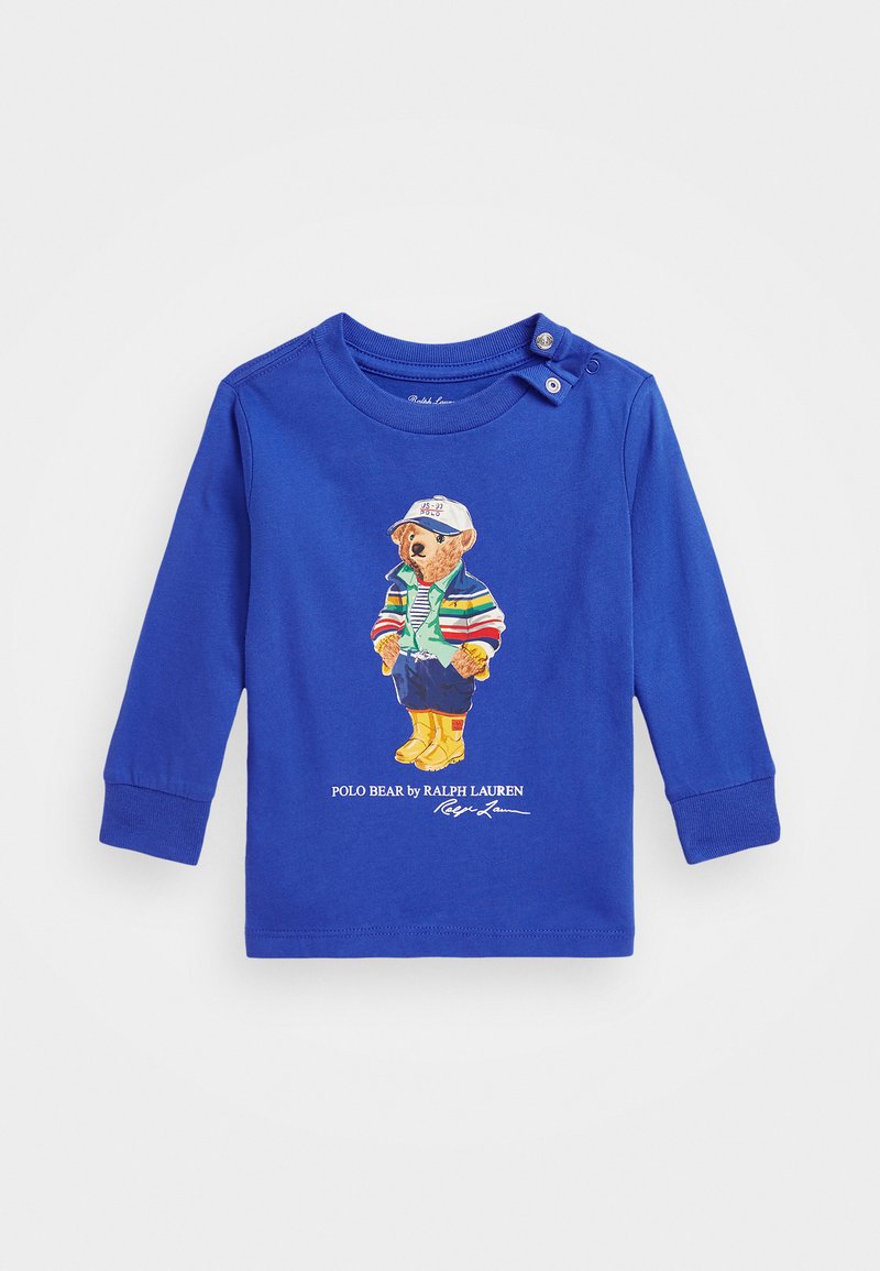 Hosszú ujjú, puha pamutból készült kék ing, színes medvével díszítve, gombos részletekkel a galléron, és nyomtatott márkanevével alul.