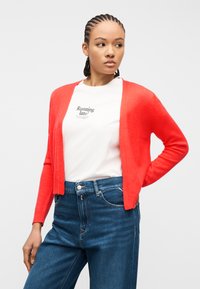 Νεαρός με πλεξούδες, φορώντας λευκό μπλουζάκι με σχέδιο, κόκκινη ζακέτα και μπλε τζιν παντελόνι, κοιτάζοντας προς τα πλευρά σε απλό φόντο.