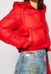 Giacca imbottita rossa con cappuccio, dotata di zip frontale, polsini elasticizzati e design corto. Abbinata a jeans blu con motivi grafici circolari.