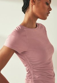 Tricou roz ajustat dintr-un material moale cu dungi, cu mâneci scurte și detalii de plisare pe laterale pentru un plus de textură. Gât rotund.