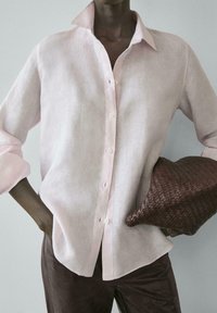 Osoba nosící světle růžovou lněnou košili na knoflíky a tmavé kalhoty, držící velké tmavě hnědé pletené psaníčko pod jednou paží.