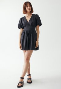 Schwarzes kurzes Kleid mit Puffärmeln, V-Ausschnitt und tailliertem Design. Der Stoff wirkt glatt mit einem gerafften Rock. Kombiniert mit schwarzen Sandalen.