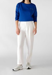 Persona vistiendo un sudadera azul con el texto "cómprate a ti mismo", pantalones blancos de cintura alta y zapatillas blancas, de pie contra un fondo sencillo.