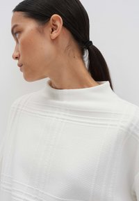 Donna con capelli scuri raccolti in una coda bassa, che indossa un maglione bianco con una texture, mentre guarda a sinistra su uno sfondo chiaro e uniforme.