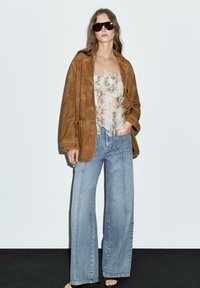 Giacca oversized in suede marrone, top chiaro in stampa serpente con volant, jeans blu a gamba larga e occhiali da sole scuri. Sfondo semplice.