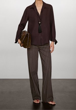 Person trägt dunkelbraunes Hemd mit Knöpfen, gestreifte weit geschnittene Hose, rote Sandalen, hält braune Wildlederhandtasche und schwarze Quasten-Halskette.