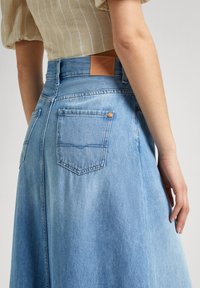 Light blue denim skirt with a mid-rise waist, back pockets, and brown leather accent at the waistband. Smooth texture and A-line shape.