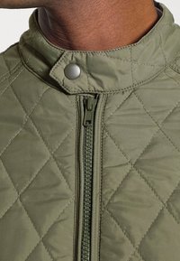 Gepolsterte olivgrüne Jacke mit hohem Kragen, silberner Druckknopfleiste und einem Frontreißverschluss mit passendem Zugband. Subtile Stickdetails.