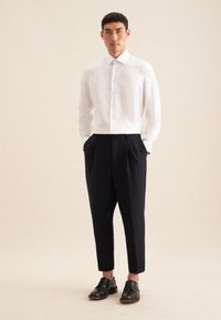 Camicia bianca a maniche lunghe con bottoni, slim fit, abbinata a pantaloni scuri a vita alta di colore blu e scarpe nere stringate. Texture morbide e linee pulite.
