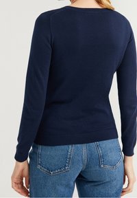 Kvinna iförd en åtsittande marinfärgad långärmad tröja och högmidjade blå jeans, sedd bakifrån.