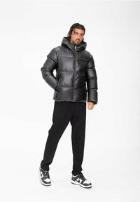 Doudoune noire avec capuche, présentant une fermeture éclair sur le devant, un design matelassé et des poches latérales. Associée à un pantalon en velours côtelé noir et des baskets.