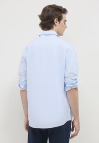 Camicia a righe blu chiaro con colletto classico, maniche lunghe arrotolate e orlo curvo. Realizzata in tessuto morbido con una texture liscia.
