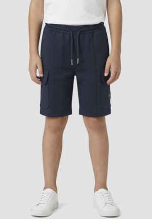 Persona con pantalones cortos cargo azul marino con cintura de cordón y zapatillas blancas, de pie frente a un fondo claro y liso.