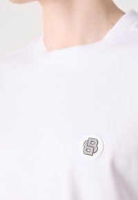 T-shirt en coton blanc avec un patch logo rond affichant un design stylisé en forme de "B" noir. Texture lisse et col rond, style minimaliste.