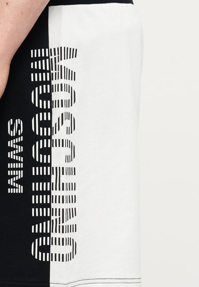 Υφασμα σε ασπρόμαυρο με το κείμενο "MOSCHINO SWIM" σε έντονα, ριγωτά γράμματα που τρέχουν κατακόρυφα κατά μήκος της διαχωριστικής γραμμής των χρωμάτων.