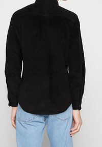 Musta corduroy-paita, jossa pystysuora ribbineulos, kauluksellinen pääntie ja napilliset kalvosimet, yhdistetty vaaleansinisiin farkkuihin.