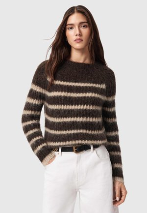 Kvinde med langt brunt hår iført en blød, mørkebrun og beige stribet sweater og hvide bukser med sort bælte, hænderne i lommerne.