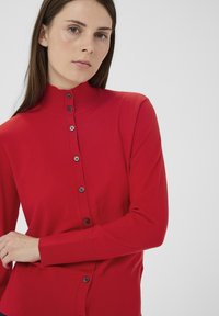 Cardigan roșu cu guler înalt. Are închidere frontală cu nasturi negri și mâneci lungi. Textură netedă, design mulat.