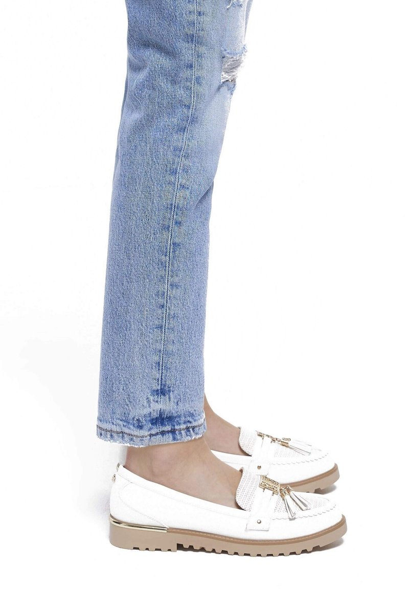Personne portant un jean déchiré bleu clair et des mocassins blancs slip-on avec des pampilles et une semelle beige, debout sur un fond blanc.