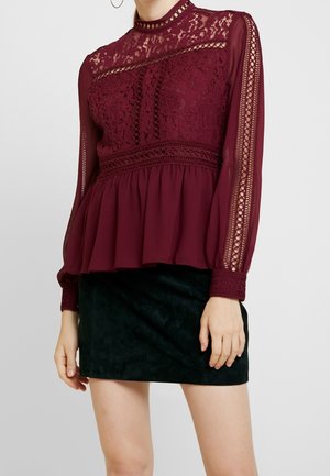 Mujer con blusa de encaje burdeos con mangas largas transparentes y mini falda negra de terciopelo, de pie frente a un fondo liso.