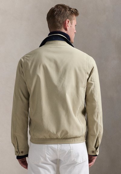 Mand med kort hår iført en beige jakke og hvide bukser, vist bagfra mod en ensfarvet grå baggrund.