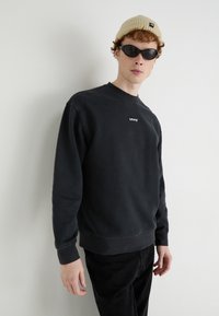 Svart sweatshirt med rund halsringning, prydd med en liten vit Levi's-logga. Matchas med svarta byxor och en beige rippad beanie.