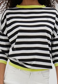 Schwarz-weiße gestreifte Bluse mit Dreiviertelärmeln, die einen neongrünen Kragen und Saum aufweist, sowie einem kleinen Herzdetail auf der Brust.