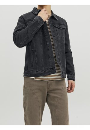 Homme portant une veste en jean noire sur une chemise beige et blanche rayée avec un pantalon marron, ajustant le col de la veste d'une main.