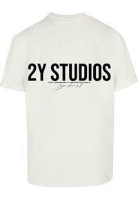 Kreemist puuvillast T-särk, millel on julged mustad kirjad "2Y STUDIOS" ja väiksem tekst "AINULT KOGUKONNA LIIKMETELE" koos kursiivis sõnaga "Syndicat."