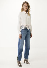 Vrouw in een witte blouse met lange mouwen en kant aan de onderkant, blauwe rechttoe-rechtaan jeans met zijsplitten en metallic heeled enkellaarzen, staande op een witte achtergrond.