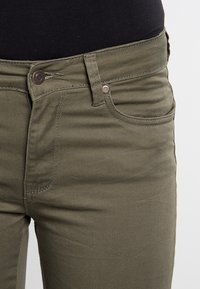 Olīvu zaļi kokvilnas maisījuma džinsi ar šauru piegriezumu, ar priekšējiem kabatām, pogu aizdari un smalkiem izšuvuma elementiem.