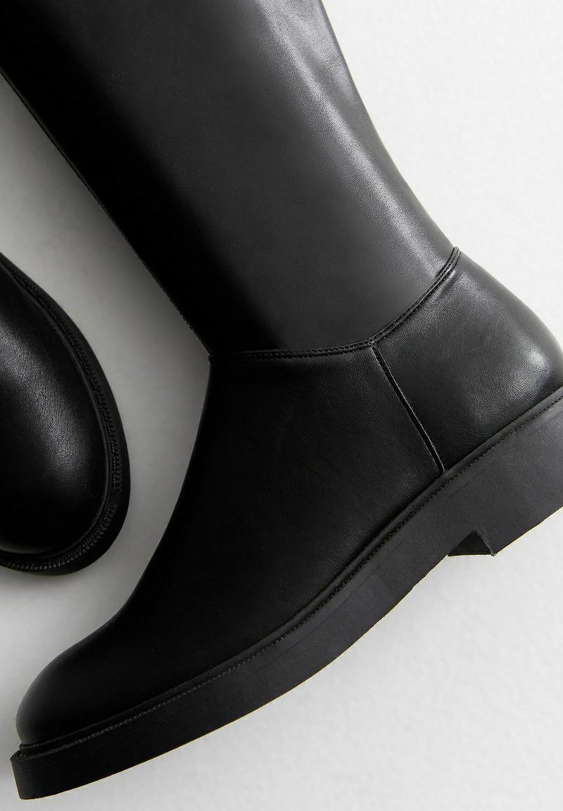 Schwarze kniehohe Stiefel aus Leder mit flacher Sohle, abgerundeter Spitze und glatter Textur. Merkmale sind minimale Nähte und ein nahtloses Design.