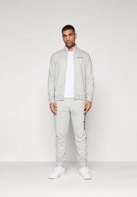 Gray zip-up sweatshirt with high collar, paired with matching jogger pants featuring a blue logo. White t-shirt and white sneakers.