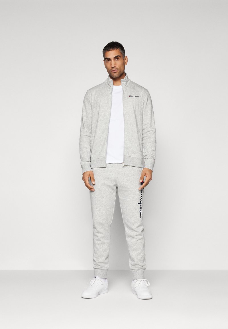 Grijze zip-up sweatshirt met hoge kraag, gecombineerd met bijpassende joggerbroeken met een blauw logo. Witte t-shirt en witte sneakers.