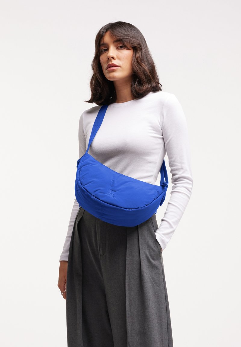 Blauw schoudertasje van zacht stof, met een afgeronde vorm en een verstelbare schouderband. Soepele textuur met een subtiele logo-details.
