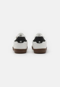 White sneakers with brown rubber soles, featuring a black heel accent. Simple design with smooth textures and a visible logo on the heel.