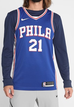 Uomo che indossa una maglia da basket blu con la scritta PHILA e il numero 21 sopra una maglietta a maniche lunghe blu navy e pantaloni corti grigio chiaro, su uno sfondo bianco.