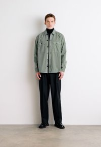 Camisa de botones verde claro con dos bolsillos en el pecho, combinada con pantalones negros y un suéter de cuello alto, vestida con zapatos negros, sobre un fondo liso.