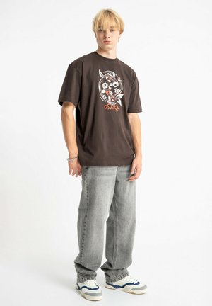 Brązowy t-shirt z grafiką przedstawiającą okrągły wzór z rybami i kwiatami. Połączony z szarymi dżinsami i trampkami z niebieskimi akcentami. Proste tło.