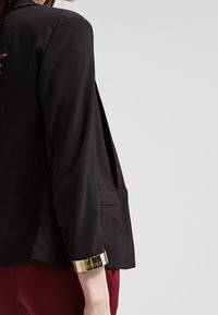 Blazer noir à manches longues, doté d'un bracelet de poignet en or. Conçu avec une coupe décontractée et des poches latérales, fabriqué dans un tissu lisse.