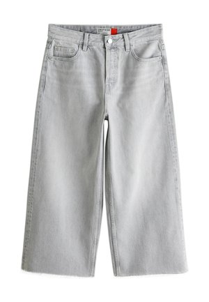 Frau mit lockigem Haar steht und trägt ein weißes kurzärmeliges Hemd mit Knöpfen, weite hellgraue kurze Jeans und flache Schuhe mit Leopardenmuster.