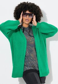 Cardigan verde lavorato a maglia con maniche sovradimensionate, indossato sopra una maglietta nera e argento con paillettes, abbinato a grandi occhiali da sole trasparenti.