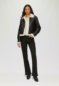 Schwarze Lederjacke mit Schafslederkragen und -manschetten, Reißverschluss, kombiniert mit engen schwarzen Hosen und schwarzen Stiefeletten mit Absatz.