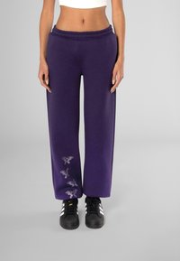 Paarse sweatpants met een elastische tailleband, versierd met decoratieve vlinderontwerpen in rhinestones langs het linkerbeen. Gekleed met zwarte sneakers.