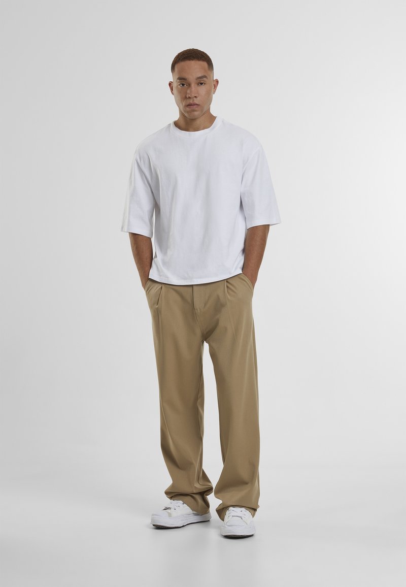 Homme debout, les mains dans les poches, portant une chemise blanche à manches courtes oversize, un pantalon beige ample et des baskets blanches sur fond gris uni.
