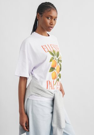 Jeune femme aux cheveux tressés portant un t-shirt blanc avec une illustration de fruit orange et un sweat-shirt gris clair noué autour de la taille.