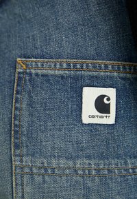 Tessuto denim di tonalità blu; presenta un piccolo etichetta bianca cucita sulla tasca con un logo nero e il nome del marchio "Carhartt."