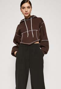 Brun cropped hettegenser med hvite snorer, kontrasterende sømmer og oversized passform, kombinert med høyhalsede svarte bukser.