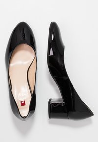 Chaussures à talons blocs en cuir verni noir avec un bout rond et une texture lisse. Elles présentent une semelle intérieure nude et un petit accent rouge sur le côté.